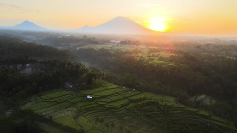 Panoramic-View-Of-Countryside-Landscape-With-Rice-Fields-And-Mountains-In-Sunset-With-Bright-Sun-In-Bali,-Indonesia---aerial-drone-shot