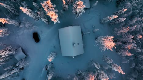 Cabin-in-middle-of-snowy-forest,-sunrise-in-Lapland---screwdriver,-Aerial-view