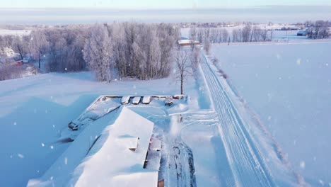 Snowing-in-rural-village-while-man-blowing-snow-near-farm-building,-aerial-view