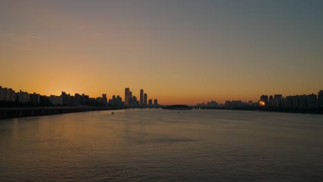 Luftaufnahme-über-Den-Fluss-Han-Und-Die-Skyline-Von-Seoul-Mit-Hohen-Gebäuden-In-Silhouettenstimmung-Zur-Orangefarbenen-Sonnenuntergangszeit