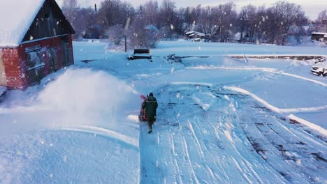 Man-plowing-snow-with-hand-machine-near-old-rural-farm-building,-aerial-view