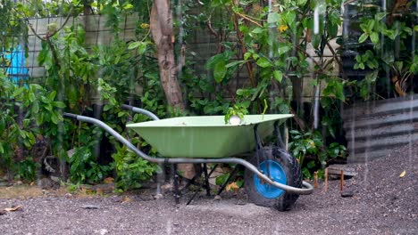 Green-wheelbarrow-resting-on-mound-of-gravel-on-building-construction-worksite-during-rain-break-caused-by-a-big-downpour-on-a-grey,-wet-day