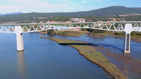 Railway-bridge-over-the-ulla-river,-the-village-and-industrial-buildings-and-the-forested-mountains-in-the-sunny-blue-sky-orizonte,-drone-shot-down,-Catoira,-Galicia,-Spain