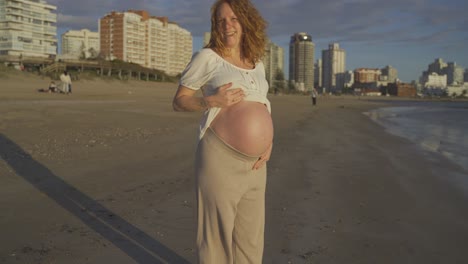 Mujer-Pelirroja-Caucásica-Embarazada-Que-Muestra-Una-Gran-Barriga-Mientras-Camina-En-La-Playa-Al-Atardecer,-Punta-Del-Este-Uruguay