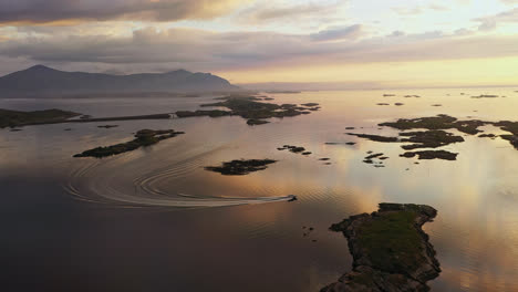 Aerial-view-following-a-boat-driving-towards-a-island-village,-with-the-Atlantic-ocean-road-in-the-background,-sunny-evening-in-Norway---tracking,-drone-shot