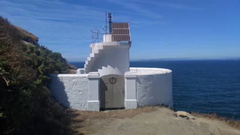 Small-white-lighthouse-with-beacon-and-solar-power-plates-on-the-edge-of-the-island-with-the-sea-in-the-background-on-a-very-sunny-afternoon,-shooting-forward