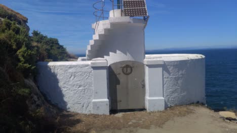 Small-white-lighthouse-with-beacon-and-solar-power-plates-on-the-edge-of-the-island-with-the-sea-in-the-background-on-a-very-sunny-afternoon,-shot-traveling-backwards