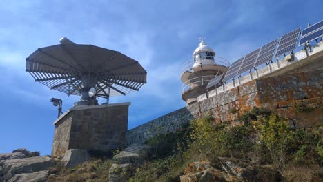Radar-for-maritime-traffic-control-with-the-lighthouse-and-solar-power-panels-at-the-top-of-the-hill-on-a-sunny-summer-day,-rolling-shot-to-the-right-from-below