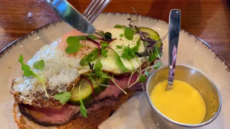 Cutting-open-a-runny-yolk-on-a-delicious-pastrami-roast-beef-sandwich-with-pickles,-microgreens-and-mustard,-breakfast-toast,-4K-shot
