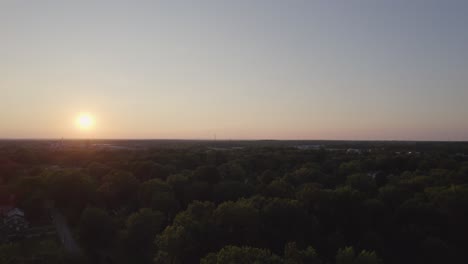 Drone-turns-back-and-captures-the-sun-setting-in-the-horizon-and-a-small-town-surrounded-by-dense-trees