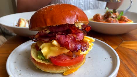 Tasty-breakfast-burger-with-eggs,-tomato,-cheddar-cheese,-bacon,-avocado-and-brioche-bun,-brunch-restaurant,-eating-in-the-background,-4K-shot