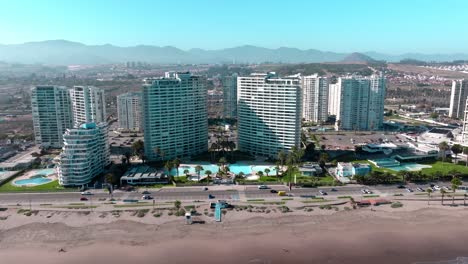Dolly-in-aerial-view-of-a-complex-of-buildings-with-swimming-pools-in-front-of-the-sea,-arid-mountains-on-the-horizon-on-a-sunny-day