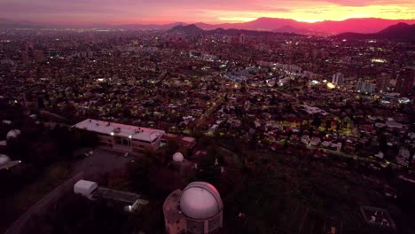 Tilt-up-aerial-view-of-the-Calan-hill-observatory-with-the-city-of-Santiago,-Chile-illuminated-at-night-with-the-sunset-in-the-background
