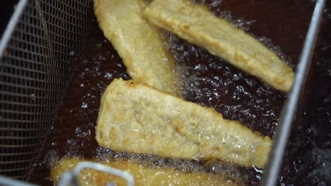 Golden-delicious-crispy-fish-fillet,-traditional-fish-and-chips-cooking-in-progress,-deep-frying-in-boiling-hot-oil-fast-food-commercial-kitchen,-night-market-pub-food,-close-up-shot
