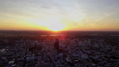 Aerial-shot-of-sunrise-in-a-city,-while-the-sun-rises-from-behind-a-building-in-a-sky-full-of-colors-and-clouds