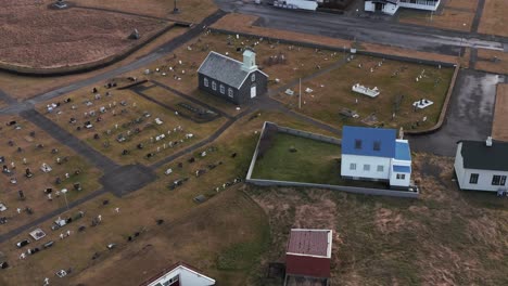 Historic-graveyard-with-church-revealing-residential-neighborhood-in-Iceland,-aerial