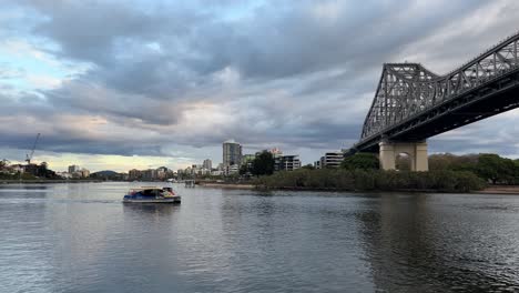 Riverside-view-capturing-a-CityCat-passenger-ferry-boat-on-the-river,-cruising-across-and-under-the-iconic-heritage-story-bridge-with-cloudy-sky-sunset,-Brisbane-City,-Queensland,-Australia