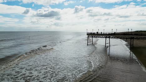 Typical-English-seaside-resort,-shot-using-a-drone-flying-into-the-sun-giving-a-aerial-viewpoint-with-a-wide-expanse-of-beach-with-a-pier-and-crashing-waves