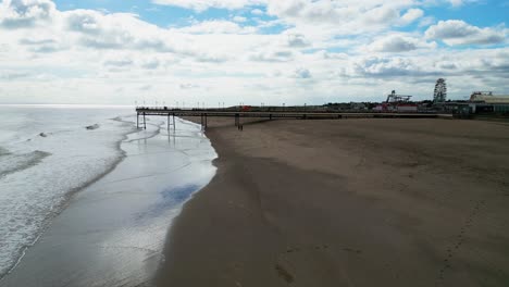 Typical-English-seaside-resort-of-Skegness,-shot-using-a-drone-flying-into-the-sun-giving-a-aerial-viewpoint-with-a-wide-expanse-of-beach-with-a-pier-and-crashing-waves