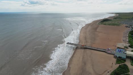 Typical-English-seaside-resort,-shot-using-a-drone,-giving-a-high-aerial-viewpoint-showing-a-wide-expanse-of-sandy-beach-with-a-pier-and-crashing-waves