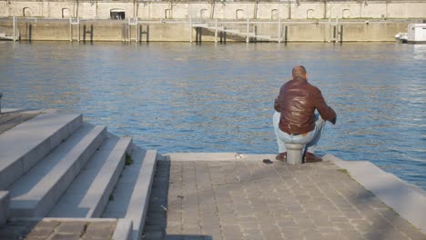 Ein-Einsamer-Mann,-Der-Auf-Dem-Dock-Einer-Bucht-Sitzt-Und-Auf-Das-Wasser-Blickt-Und-Kaffee-Trinkt