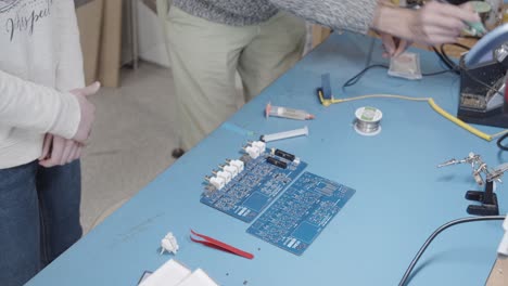 Man-and-woman-working-in-electronics-laboratory-welding-blue-electronic-board-with-welder-and-tin-while-wearing-COVID-19-masks