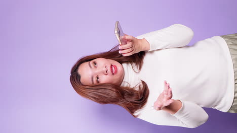 Vertical-of-Young-Asian-women-talk-on-a-phone-using-loudspeaker-on-technology-to-network-with-clients-or-negotiate-deals-isolated-over-a-violet-background-1