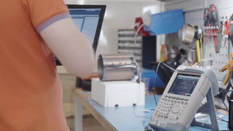 Electronics-engineer-wearing-a-COVID-19-mask-while-programming-software-and-holding-the-laptop-in-his-arm-working-with-spectrum-analyzer-in-electronics-laboratory