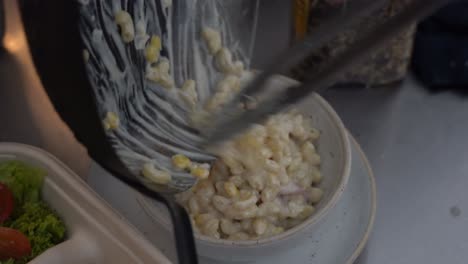 Professional-chef-pouring-and-scraping-cooked-macaroni-and-cheese-into-a-bowl,-delicious-and-tasty-adults-and-kids-favourite-comfort-food-dish,-handheld-motion-close-up-shot