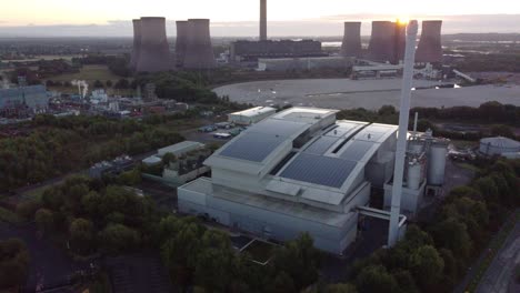 Die-Luftaufnahme-Senkt-Sich-über-Eine-Intelligente-Solarfabrik-Auf-Dem-Dach,-Wobei-Der-Sonnenaufgang-Hinter-Dem-Kraftwerk-Für-Fossile-Brennstoffe-Auftaucht