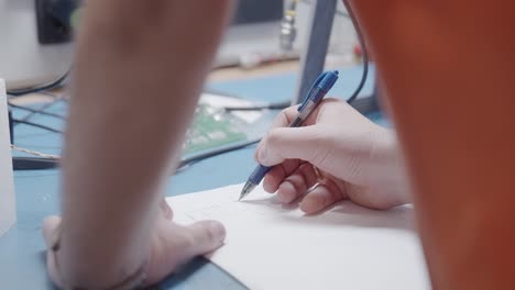 Electronics-engineer-drawing-electronics-circuit-plan-with-a-blue-pen-over-a-blue-workbench-with-cables-and-motherboard-in-the-background