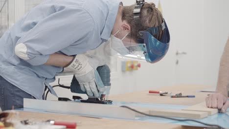 Man-working-wearing-a-covid-mask,-protection-gloves-and-face-shield-cutting-metal-with-electric-jigsaw-while-another-man-holds-the-metal-piece