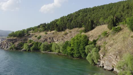 Drone-flying-from-Lake-Ohrid-to-gree-tree-filled-coastal-cliffs,-North-Macedonia