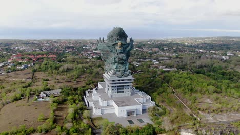 Massive-statue-of-Garuda-Winu-Kencana-near-small-town-of-Bali-island,-aerial-orbit-view