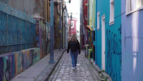 Curly-haired-blonde-tourist-girl-walking-with-her-back-to-the-camera-through-a-graffitied-urban-alley-in-Valparaiso,-Chile