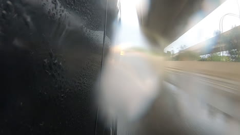 Rain-running-down-on-camera-as-a-black-car-drives-along-a-highway-during-a-rainy-late-afternoon-with-the-camera-facing-the-front-wheel-and-on-coming-traffic-under-road-bridges