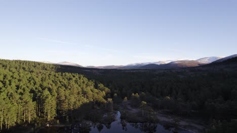 Aerial-drone-footage-over-the-surface-of-still-water-with-reflections-in-the-Cairngorms-National-Park,-Scotland-revealing-a-native-scots-pine-forest-and-snow-capped-mountain-landscape