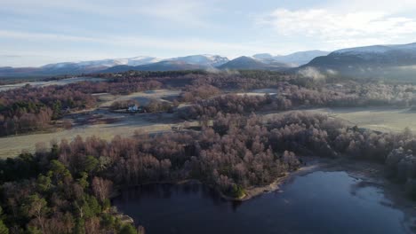 Aerial-drone-footage-of-frost-covered-trees-and-forest,-snow-capped-mountains-and-frozen-loch-in-a-frozen-winter-landscape-on-Rothiemurchus-estate-in-Cairngorms-National-Park,-Scotland