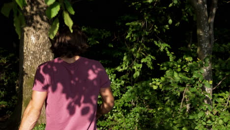 Young-man-in-red-T-Shirt-and-long-hair-walks-in-slowmotion-into-a-green-dark-forrest-on-a-sunny-summer-Day