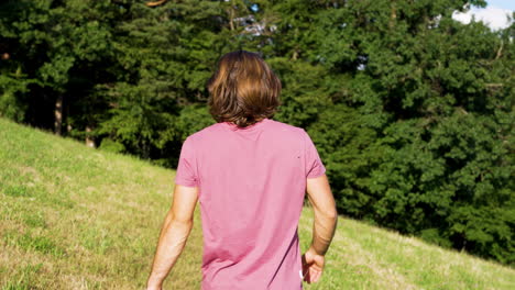 Young-man-in-red-T-Shirt-and-long-hair-walks-in-slowmotion-on-a-green-meadow-towards-a-forrest-on-a-sunny-summer-Day