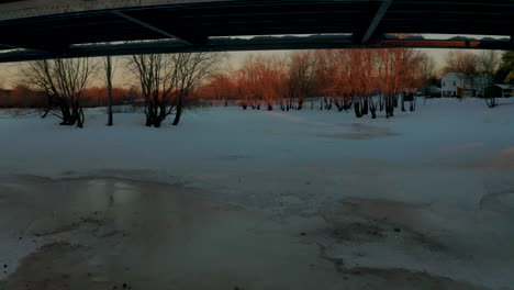 Aerial-view-flying-under-a-bridge-and-along-a-frozen,-snowy-river-during-a-scenic-winter-sunset
