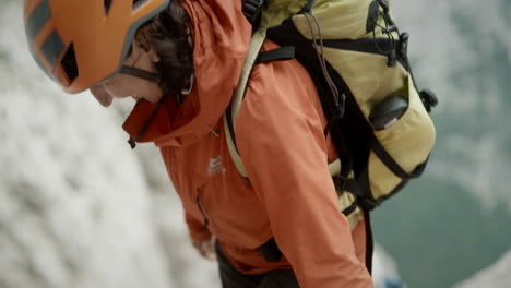 A-happy-hiker-climbing-up-a-mountain-wearing-orange-jacket-and-a-helmet-and-a-backpack,-up-close