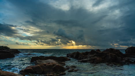 Golden-god-rays-break-through-clouds-at-sunset-over-ocean---coastal-time-lapse