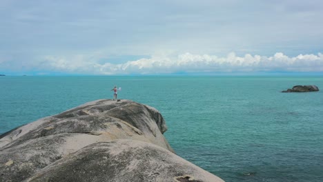 Niña-Corriendo-Sobre-Grandes-Rocas-De-Granito-Hacia-El-Océano-Azul-Turquesa-En-Pantai-Penyabong-En-Belitung-Indonesia-En-Un-Día-Nublado,-Antena