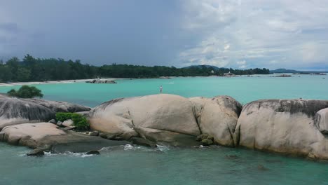 girl-standing-on-large-granite-boulders-with-turquoise-blue-ocean-on-cloudy-day-in-Pantai-Penyabong-Belitung,-aerial