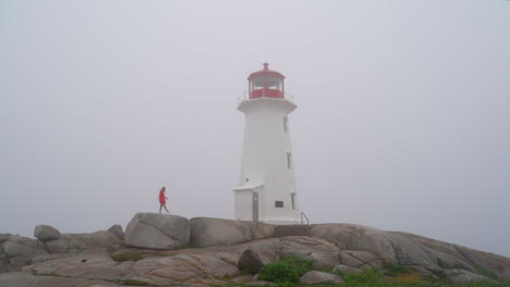 Young-woman-walking-with-a-lighthouse-in-the-fog-on-an-overcast-day-in-Nova-Scotia,-Canada-Peggy-Cove