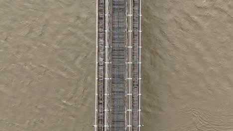 Top-Down-Aerial-View-of-Old-Suspension-Bridge-Above-Flooding-Muddy-River