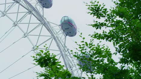 Nahaufnahme-Des-London-Eye,-Das-Sich-Langsam-An-Einem-Bewölkten-Tag-Dreht,-Mit-Grünen-Baumblättern,-Die-Sich-Im-Wind-Bewegen