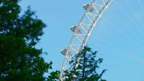 Lockdown-in-London,-smooth-slow-motion-pan-of-the-static-London-Eye-against-green-trees-and-sunny-blue-skies,-during-the-Coronavirus-2020-pandemic