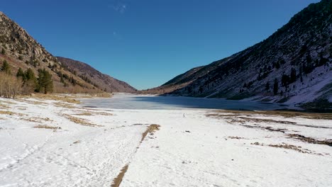 Antena-De-Una-Mujer-Y-Su-Perro-Caminando-Junto-A-Un-Lago-Helado-De-Invierno-En-Las-Montañas-De-Sierra-Nevada,-Cerca-De-Yosemite,-California